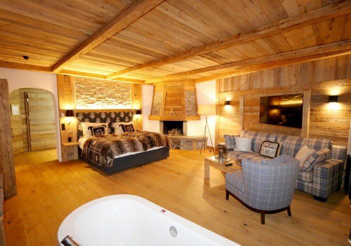 Cozy rustic bedroom with wood paneling, a bed, fireplace, armchairs, and a jacuzzi tub in the foreground