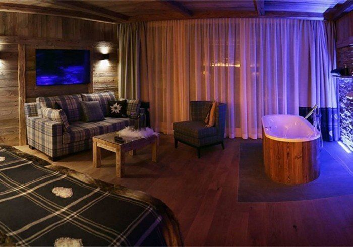 Cozy wood-paneled lounge with plaid sofa, TV, coffee table, chair, and glowing bathtub by sheer curtains.