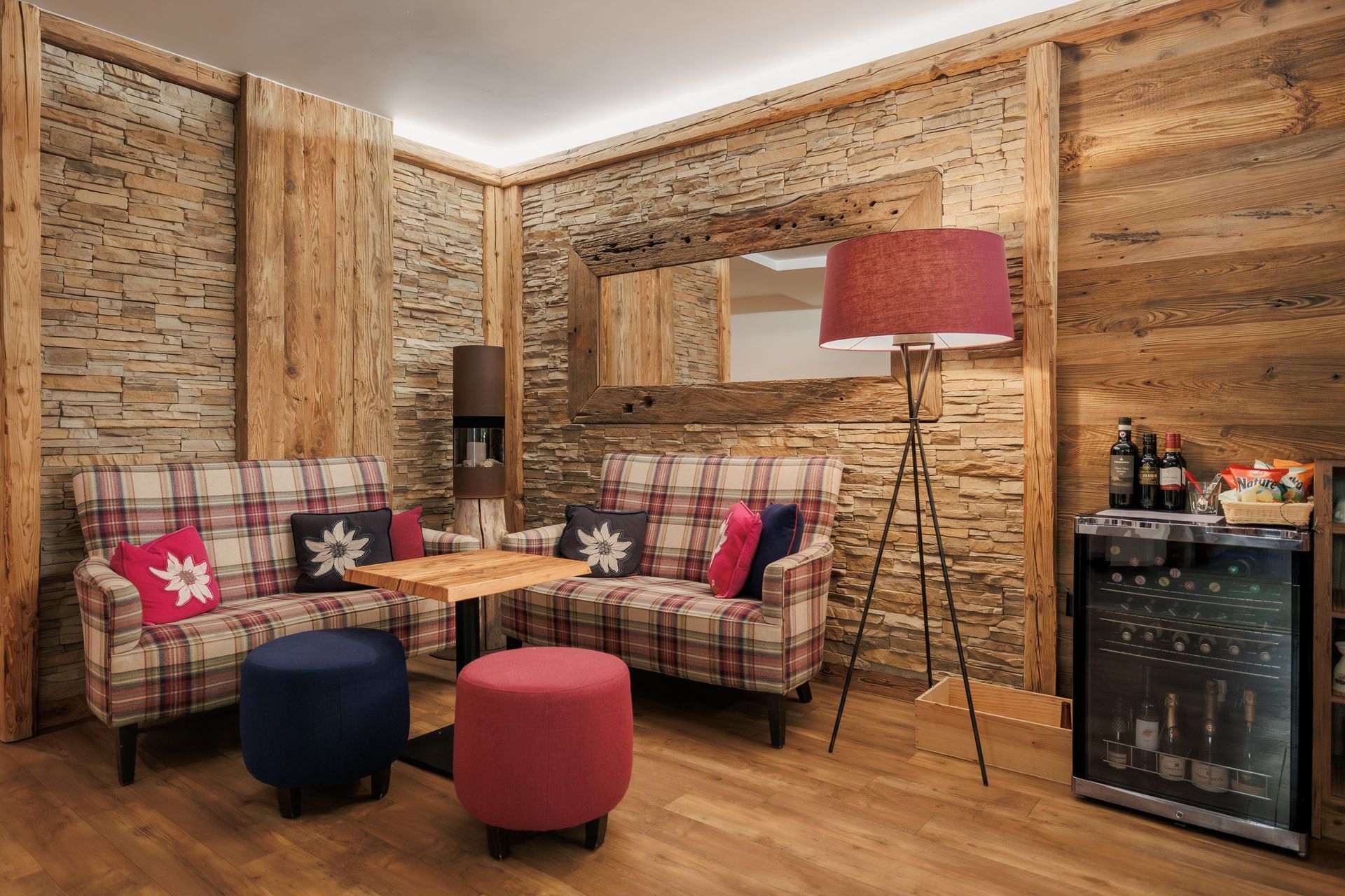 Cozy rustic lounge with plaid sofas, colorful ottomans, floor lamp, and a mini fridge against wood-paneled walls