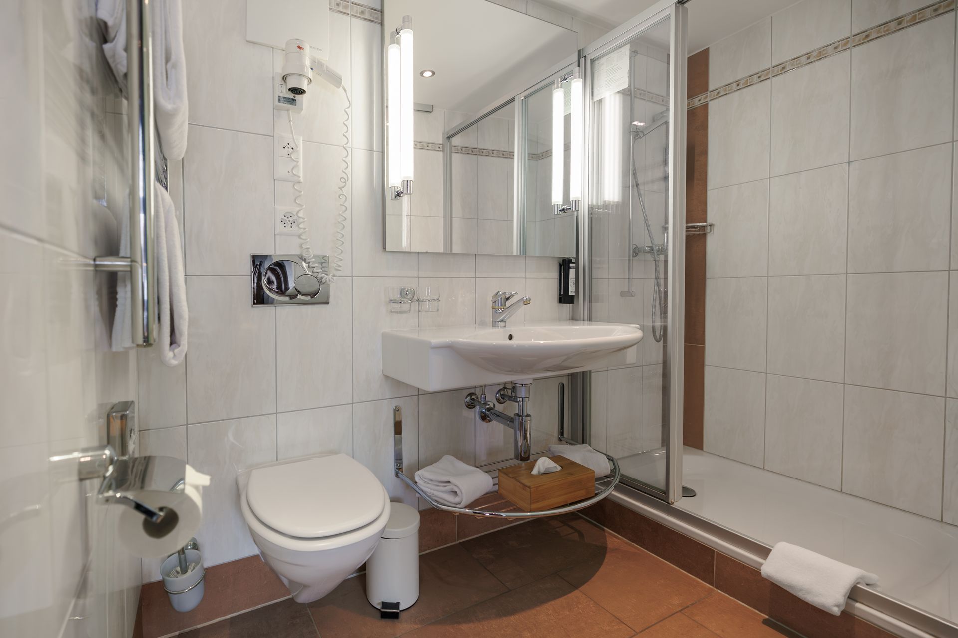 Small tiled accessible bathroom with toilet, sink, mirror, shower, and grab bars