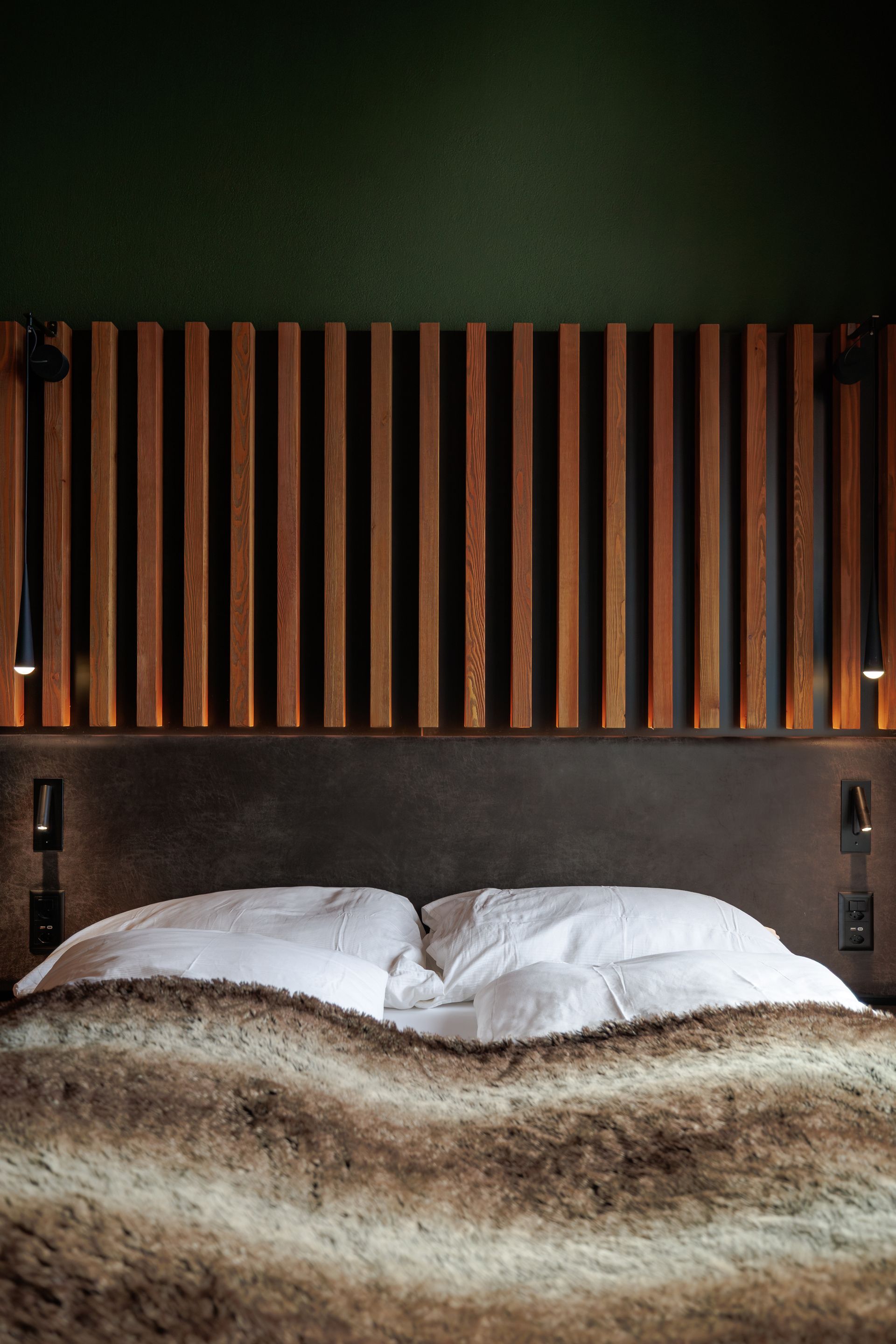 Bed with white pillows and striped blanket beneath a wooden headboard in a dark green bedroom.