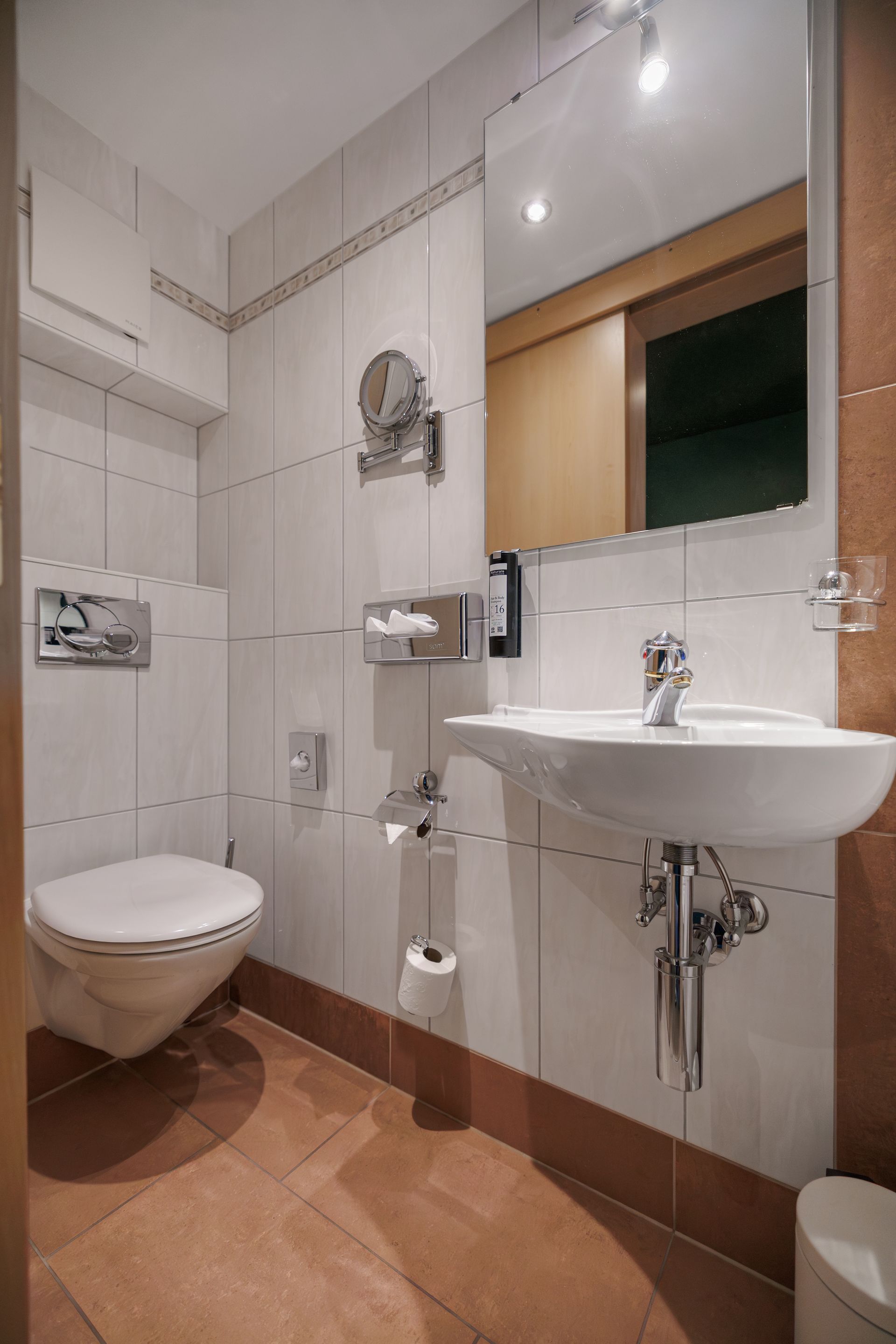 Small tiled bathroom with wall-mounted toilet, sink, mirror, and shower corner