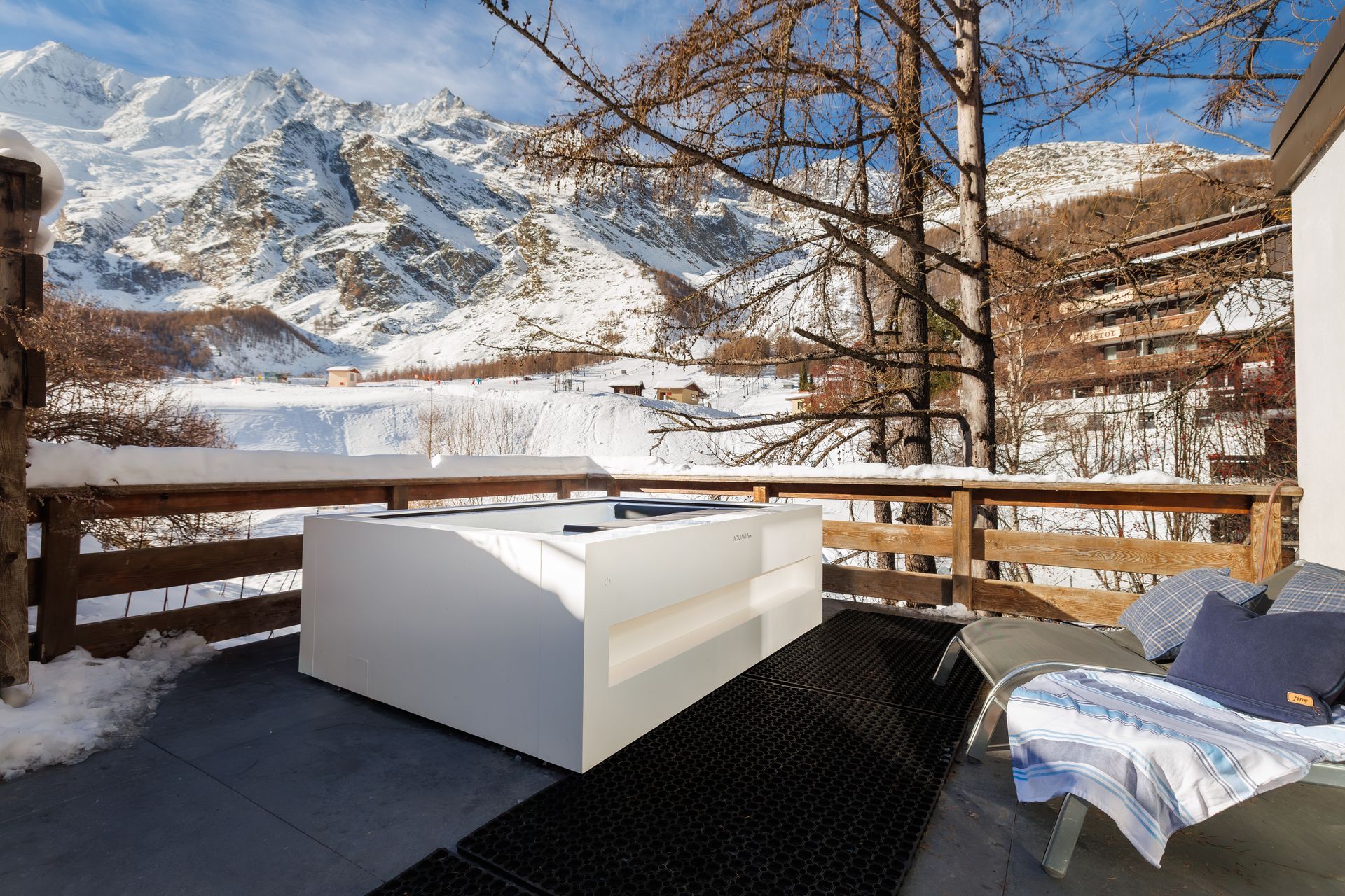 Snowy mountain-view balcony with white outdoor sofa and table, wooden railing, and leafless trees