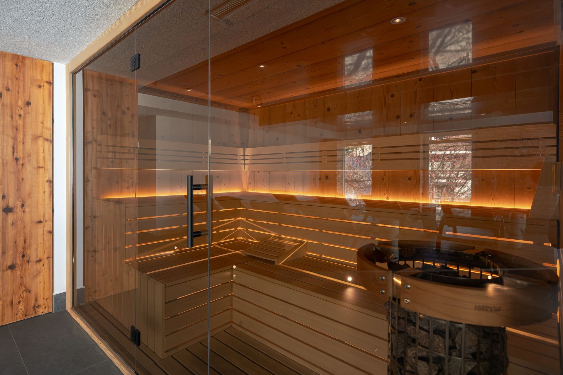 Modern sauna with warm wood panels, glass walls, and amber lighting, featuring benches and a round heater.