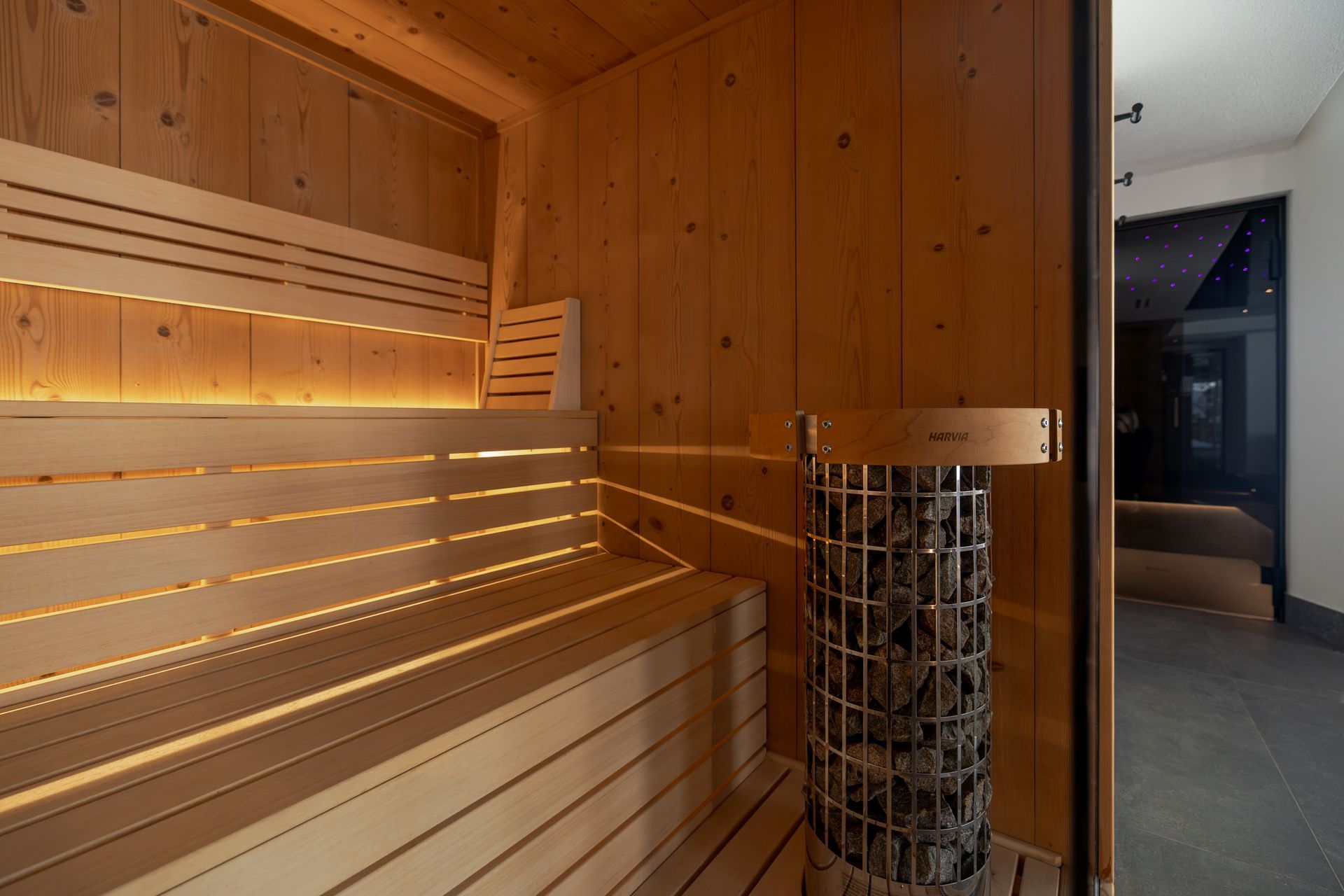 Wood-paneled infrared sauna with glowing bench lights and a glass door to a hallway