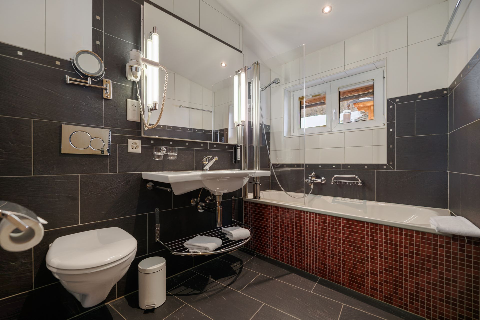 Modern bathroom with black tile, white sink and toilet, and a bathtub by a window
