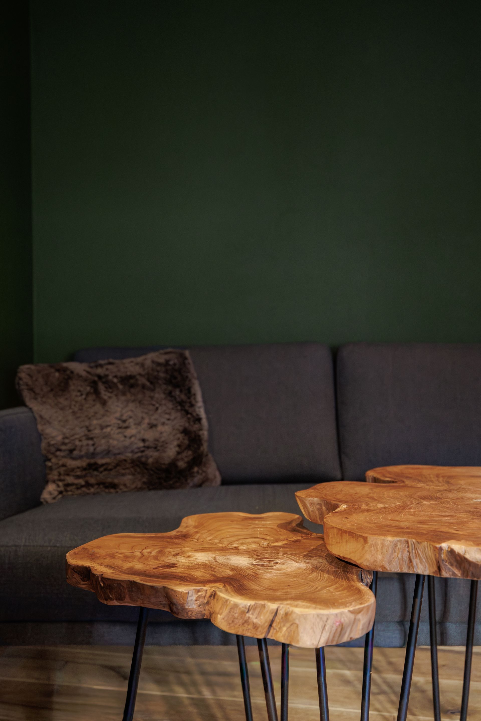 Dark green lounge with a gray sofa, fur pillow, and two rustic wooden coffee tables on black metal legs.