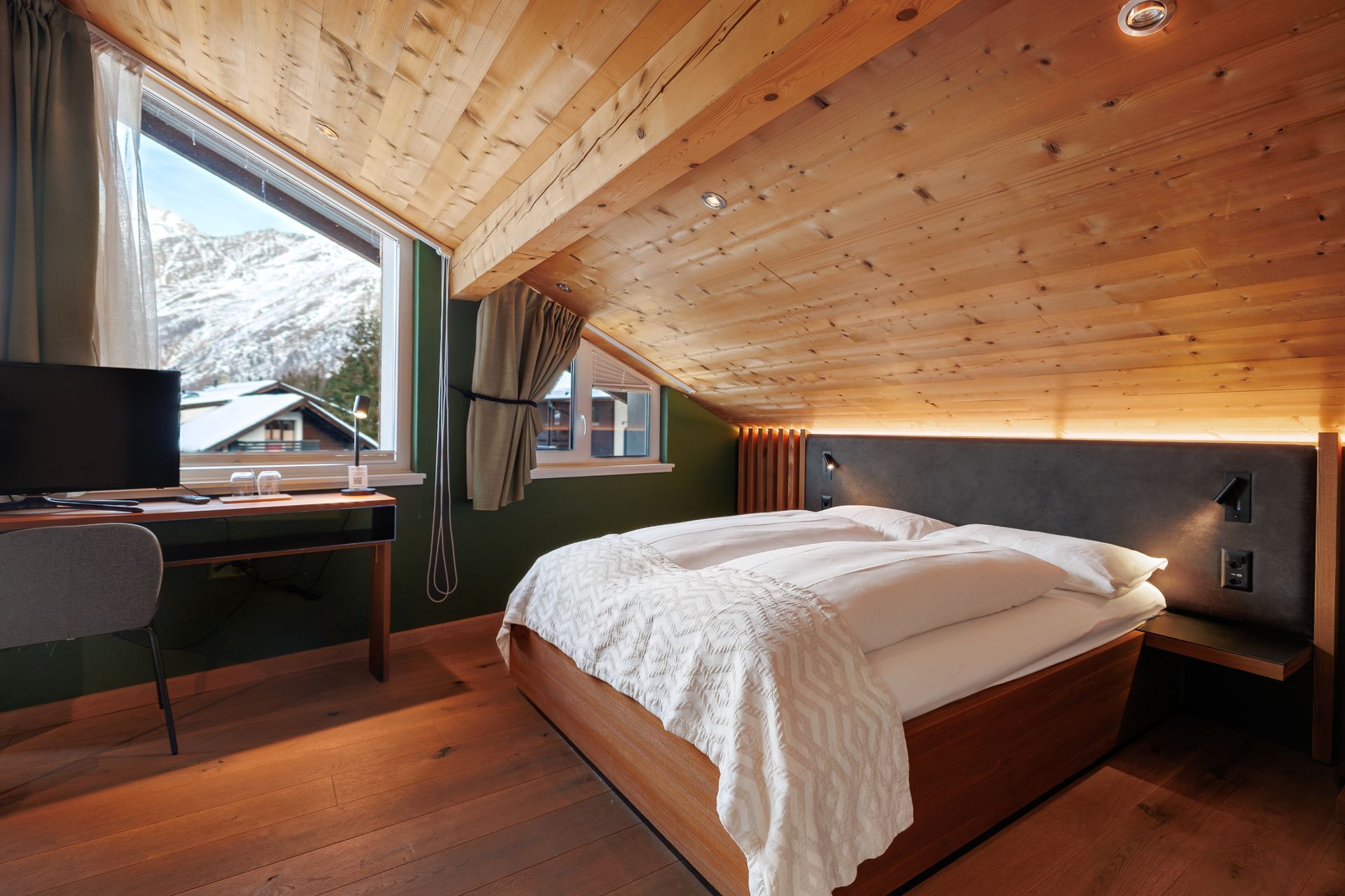 Cozy attic bedroom with sloped wood ceiling, bed, desk, and windows overlooking trees