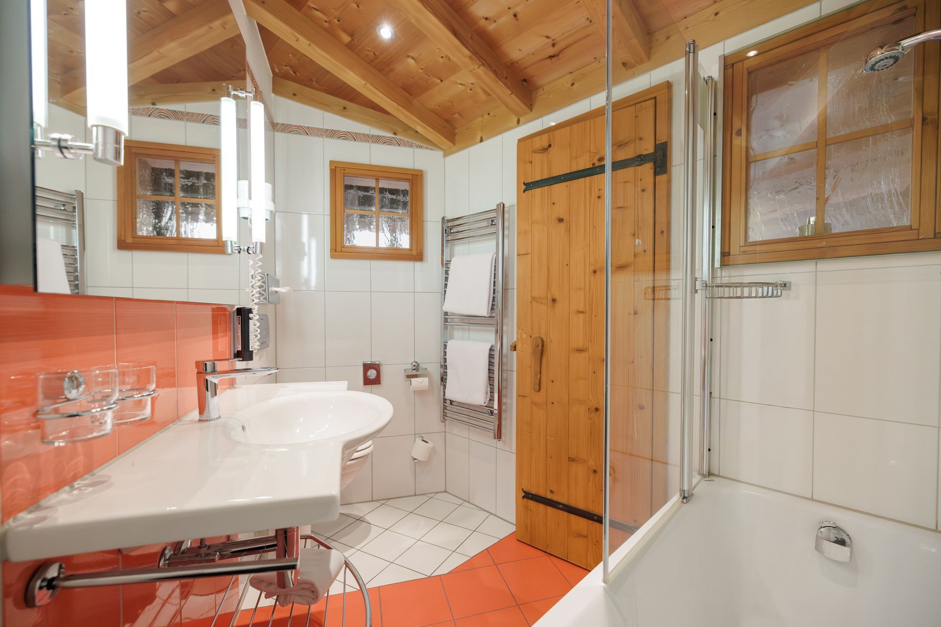 Bright bathroom with orange tile, white sink and tub, wooden ceiling, and glass shower enclosure