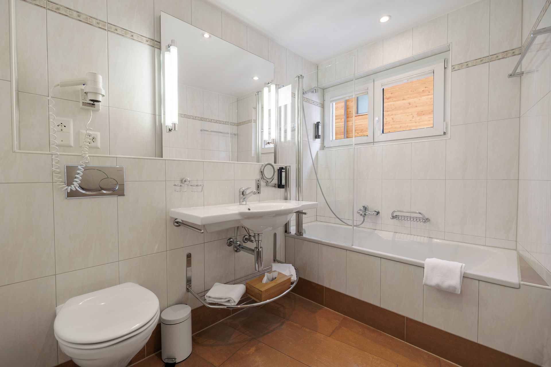 Bright white bathroom with toilet, sink, bathtub, and mirror, with brown tiled floor and towels hanging.