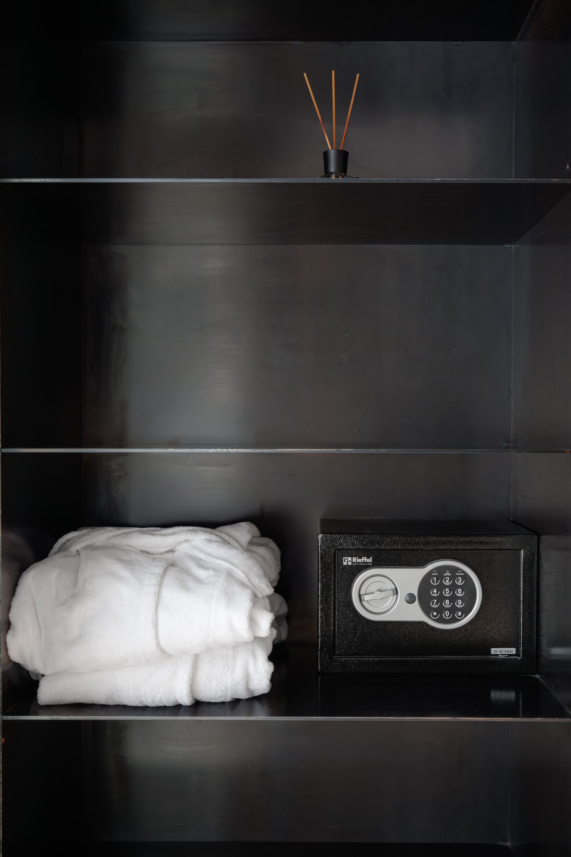 Black shelf with folded white towels, a small safe, and reed diffuser on the top shelf.