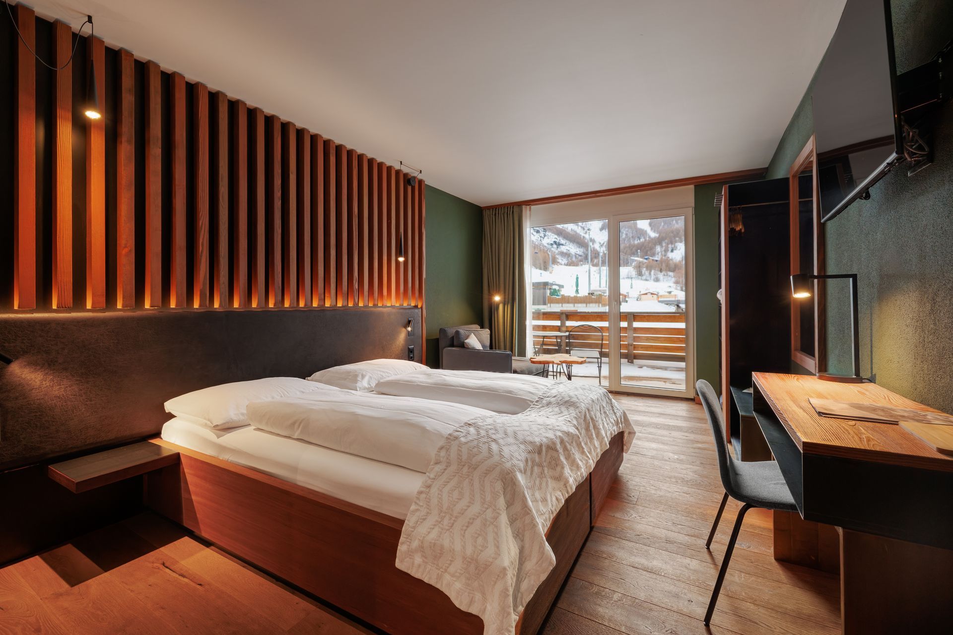 Modern hotel room with a large bed, wooden slat wall, and balcony view of snowy mountains
