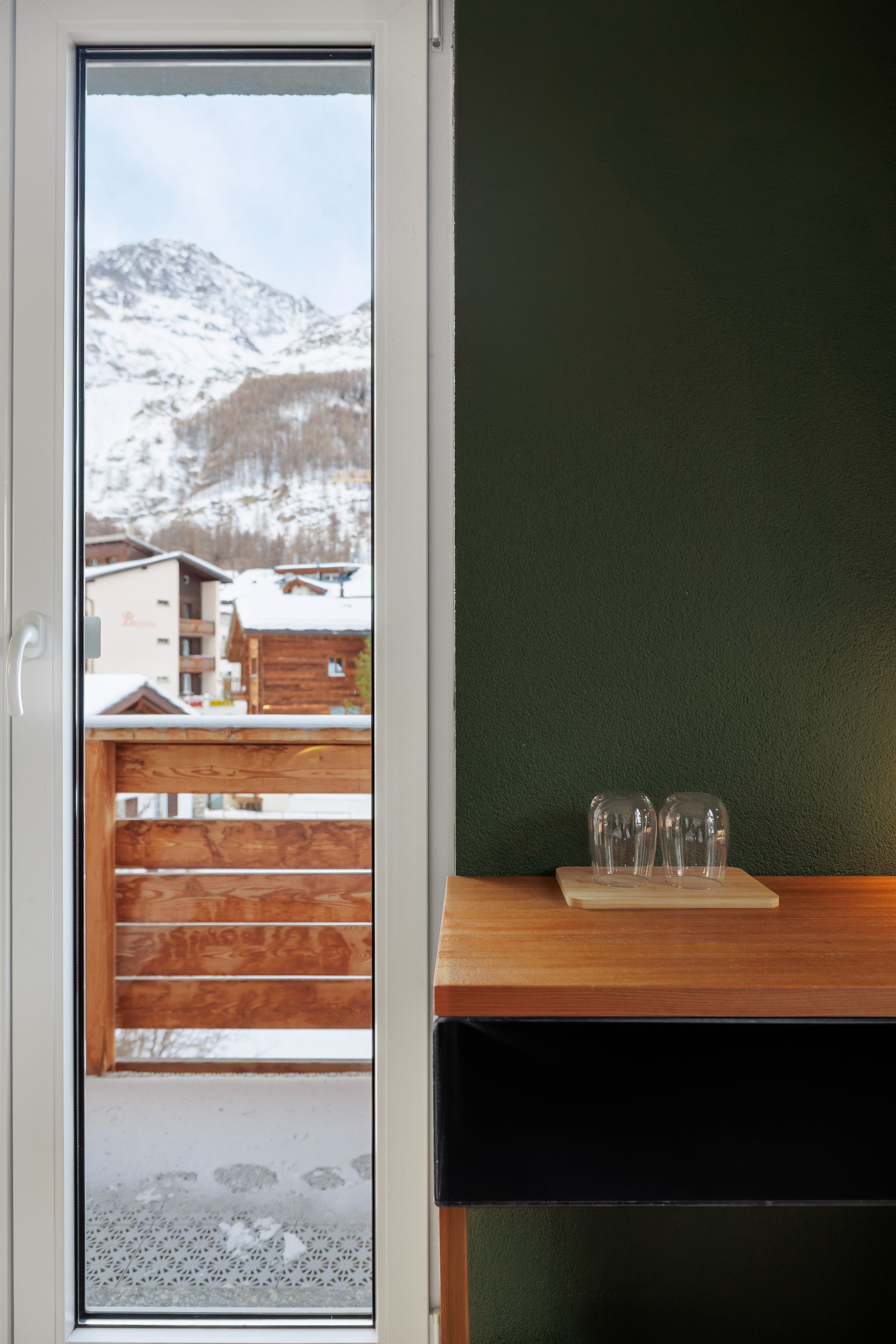 Interior doorway with snowy mountain view outside, wooden balcony railing, and a small table against a dark green wall