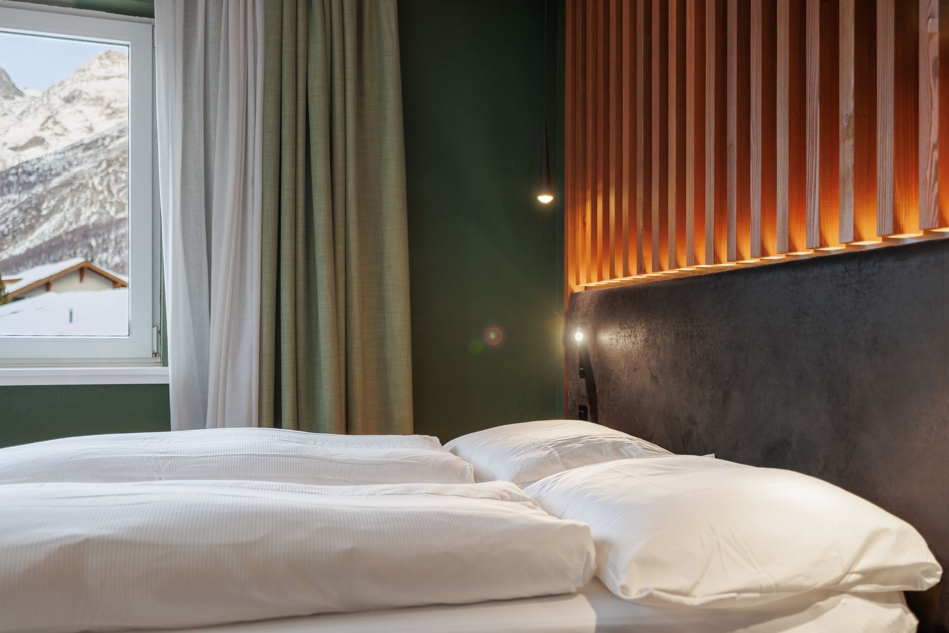 Cozy bedroom with white bedding, dark headboard, warm wall lights, and snowy mountain view outside the window