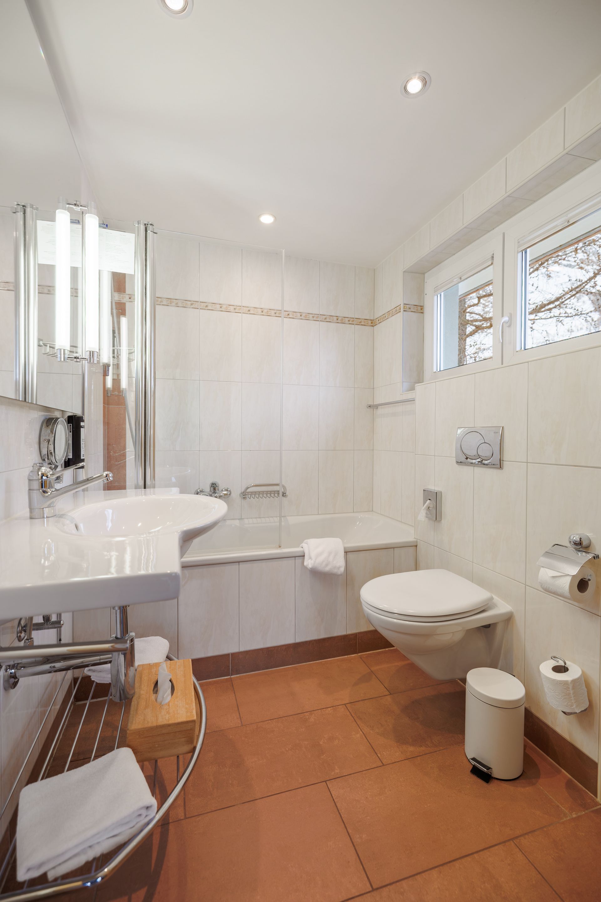Bright white bathroom with bathtub, sink, toilet, terracotta tile floor, and window light