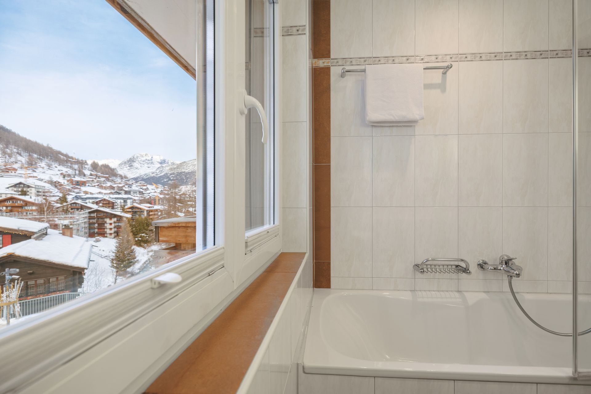 Bright bathroom with bathtub and shower beside a large window overlooking snowy houses.