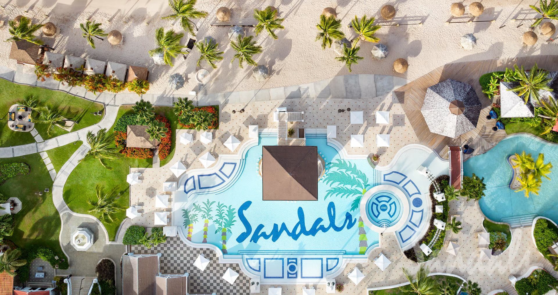 Aerial view of a sandy resort pool with blue “Sandals” logo, palm trees, and lounge chairs