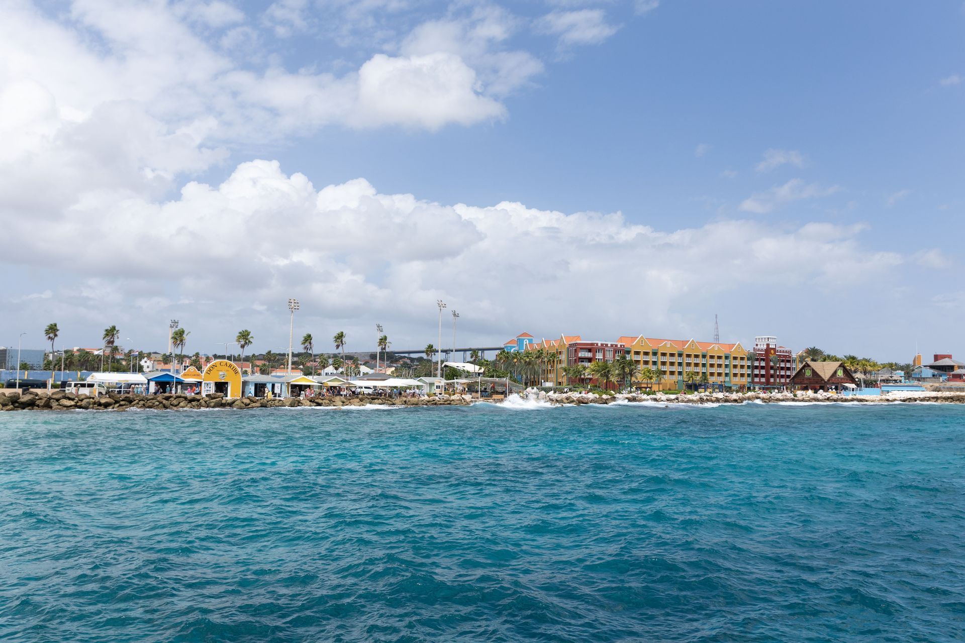 Coastal resort with colorful buildings, palm trees, and turquoise ocean under a partly cloudy sky