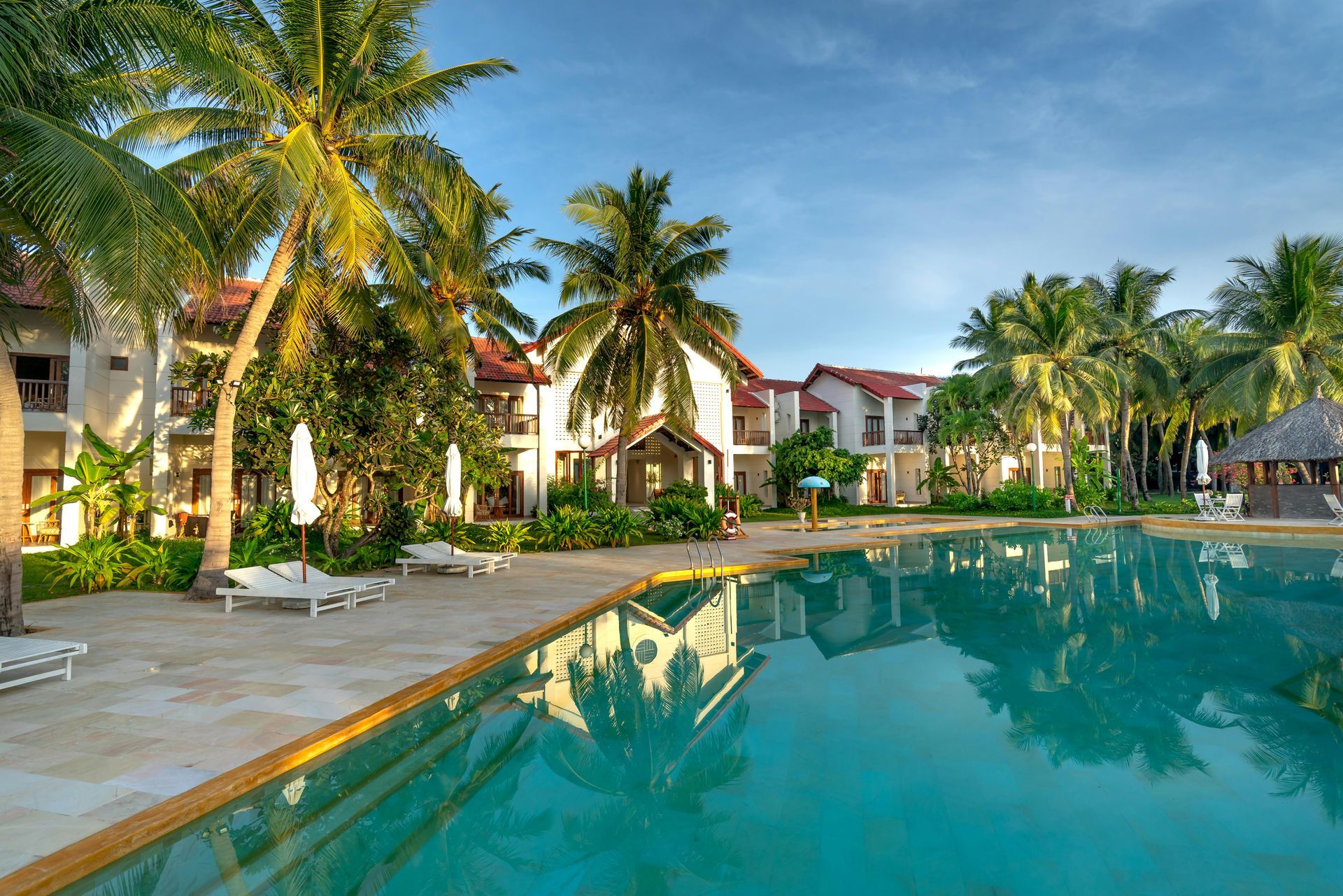 Palm-lined resort pool with turquoise water, lounge chairs, and villa buildings under a clear sky