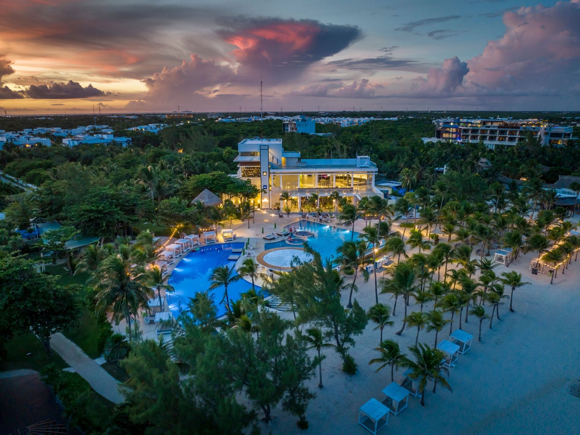 Aerial view of a tropical resort with lit pool, palm trees, and beach at sunset under a colorful sky