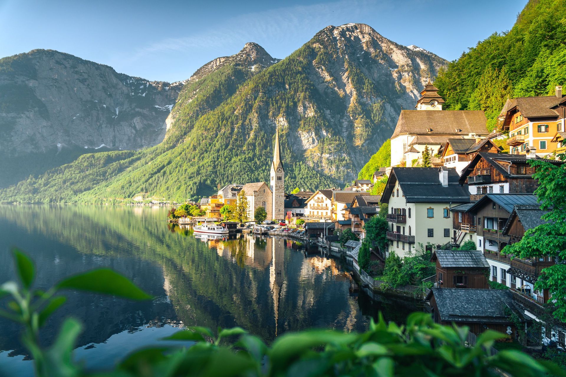 Mountain village beside a calm lake, with sunlit houses, green hills, and rugged peaks reflected in the water
