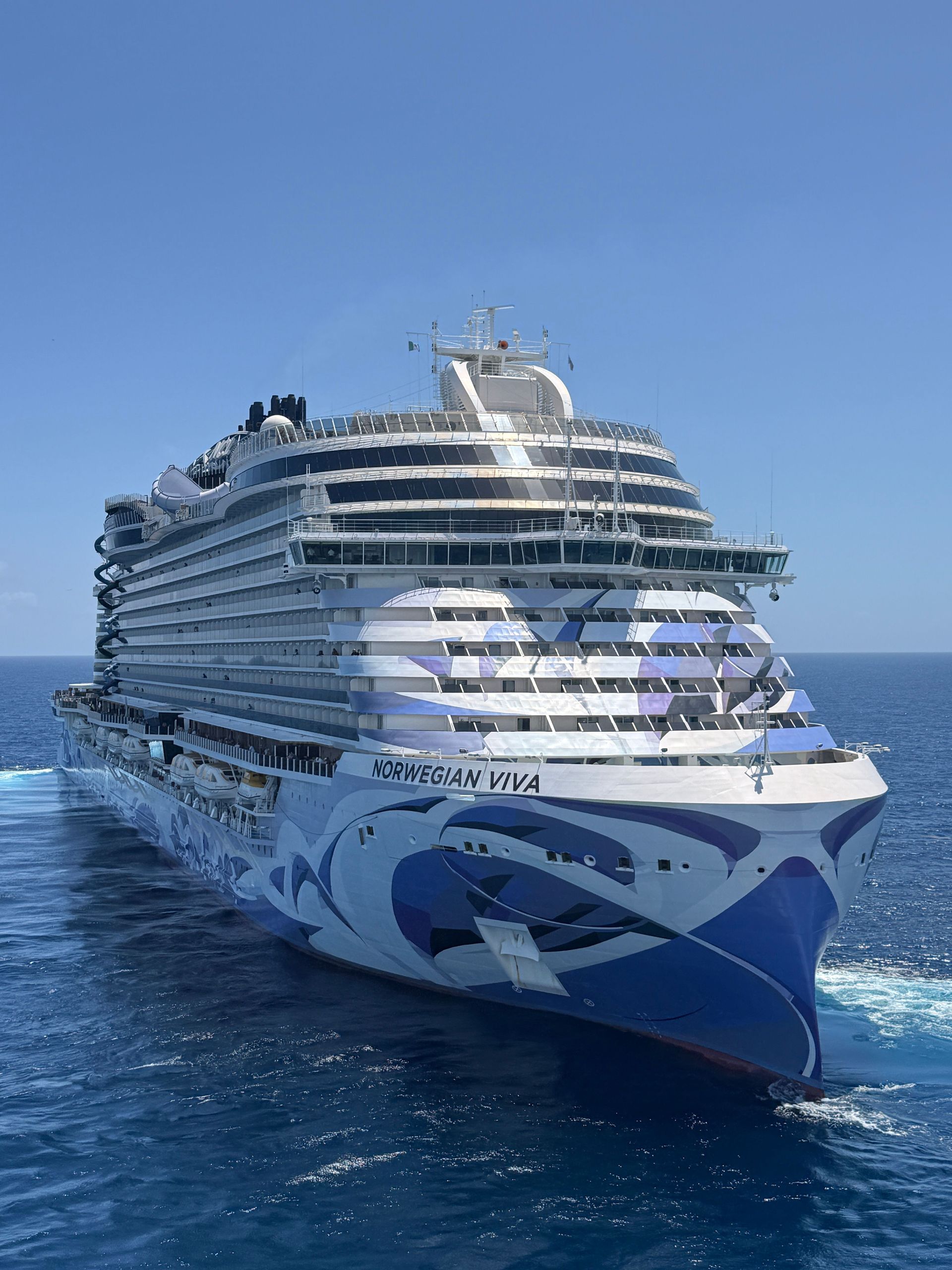 Large white cruise ship sailing on calm blue water, seen from above.