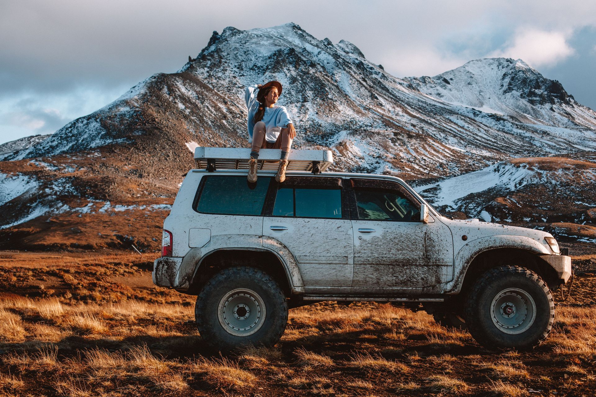 Mud-covered white SUV parked in a snowy mountain landscape with a person sitting on the roof rack