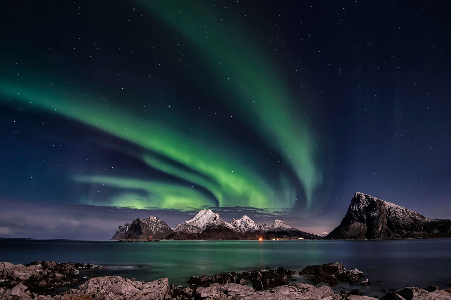 Northern lights ripple over snowy mountains and a calm sea at night.