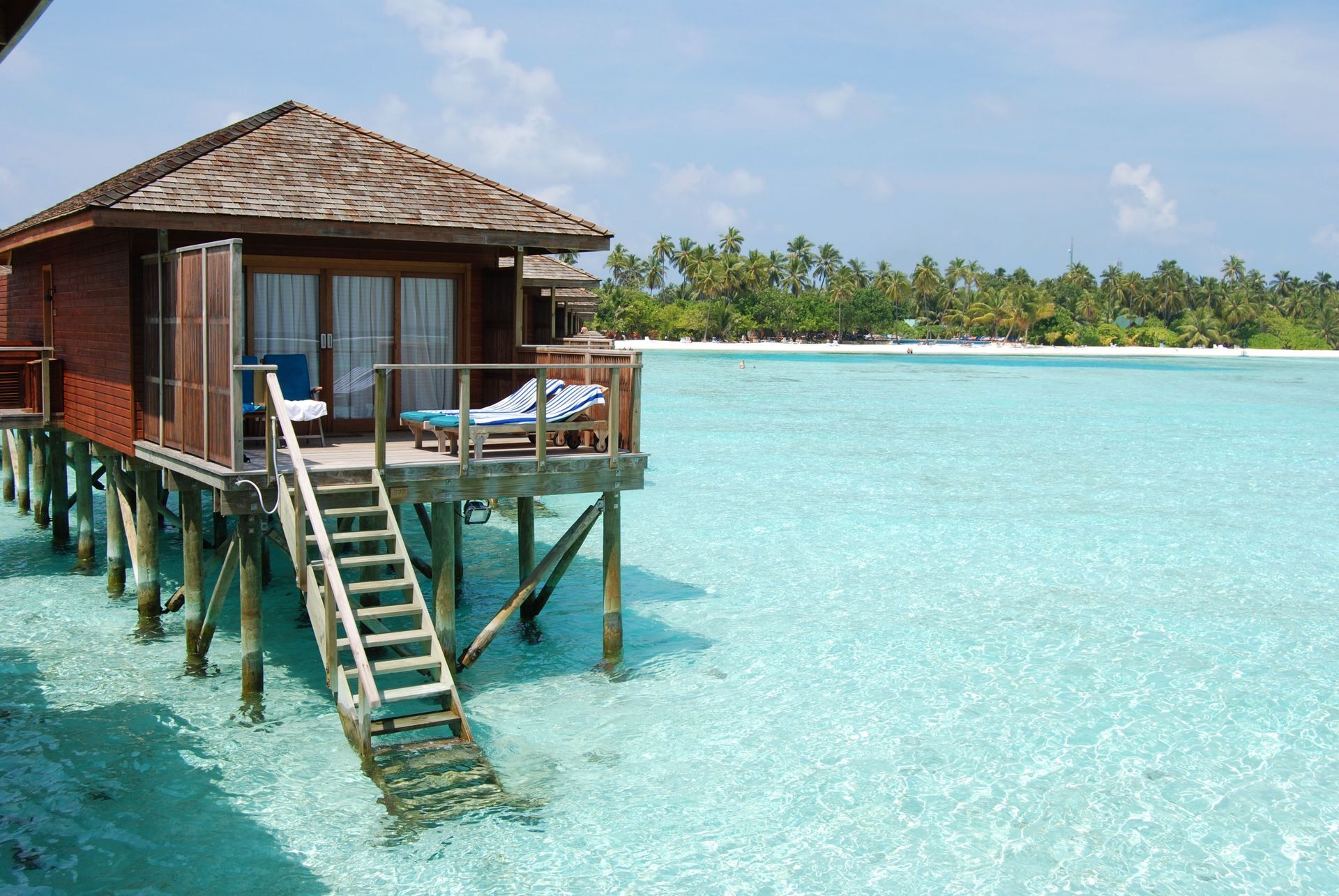 Wooden overwater bungalow with stairs above clear turquoise lagoon and palm-lined shore
