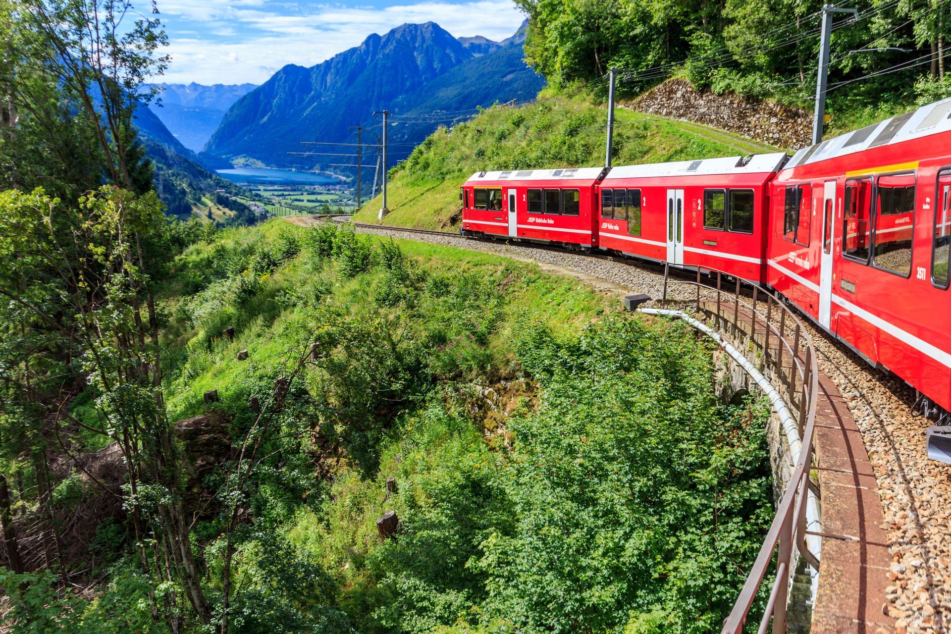 Red mountain train curving through lush green hills with snowy peaks in the distance