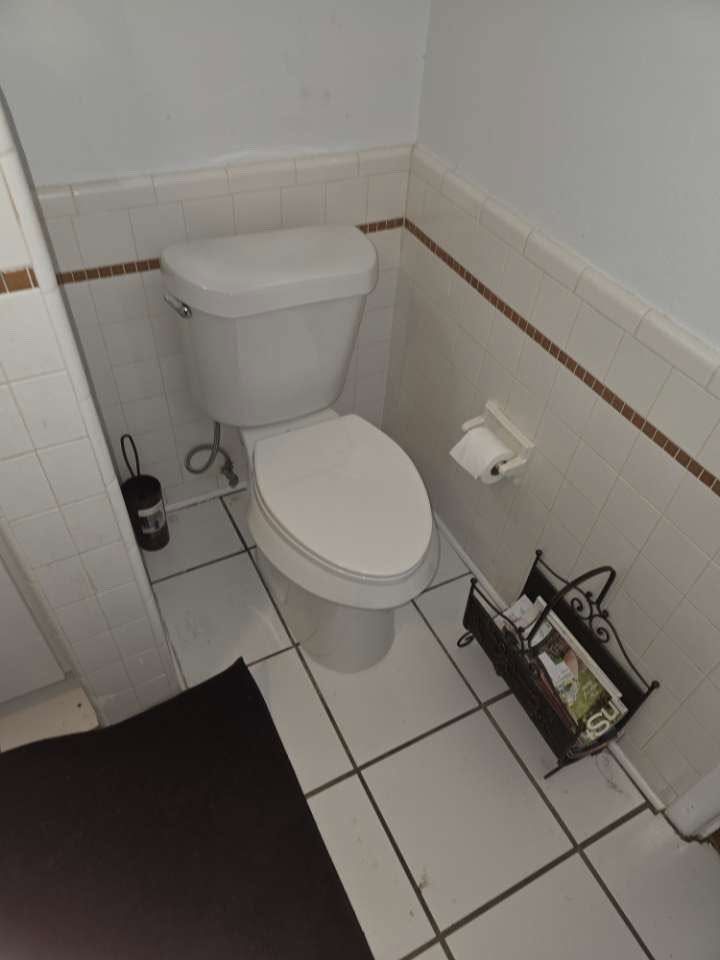 Small tiled bathroom with a white toilet, toilet paper holder, and black floor mat.