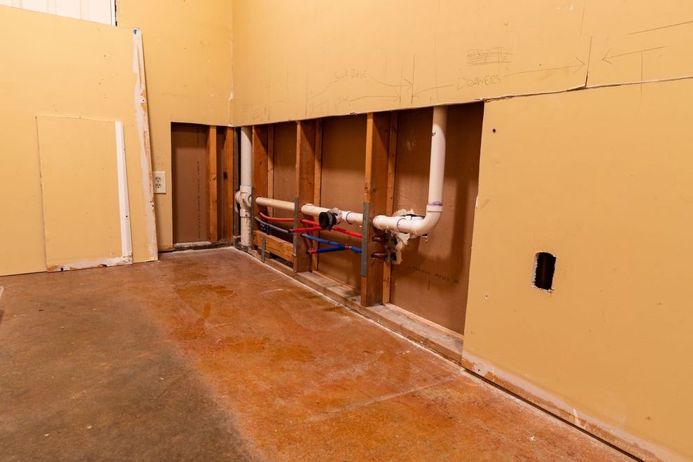 Damaged interior wall with exposed pipes and open studs in an unfinished room