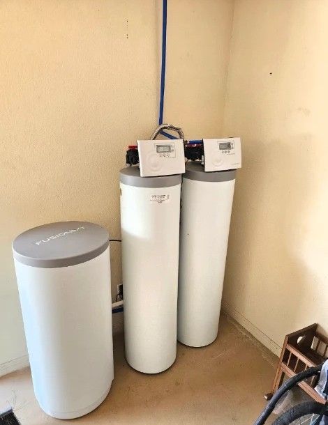 Three white water softener tanks in a utility corner with a blue pipe overhead.