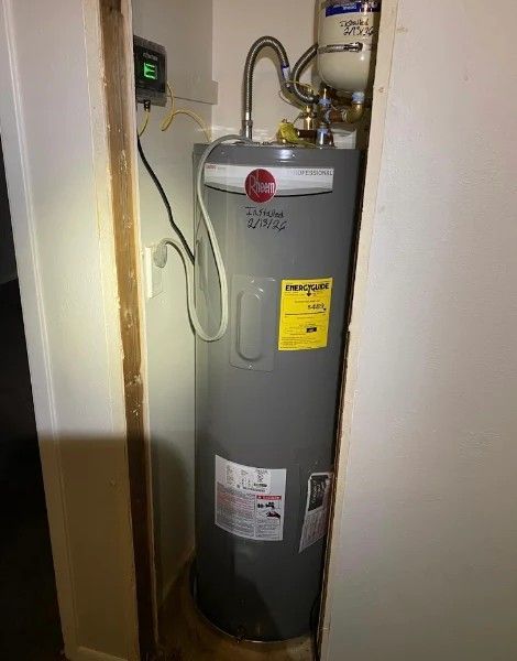 Gray water heater in a narrow utility closet with pipes, valves, and a wall control panel.