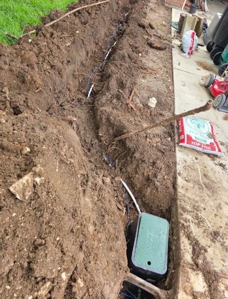 A narrow trench with exposed dirt and cables beside a concrete walkway and garden tools.