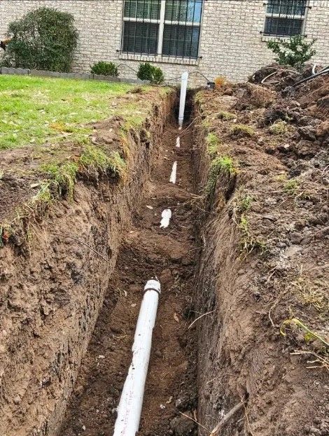 A deep trench with exposed white PVC pipes runs beside a house foundation and grassy yard.