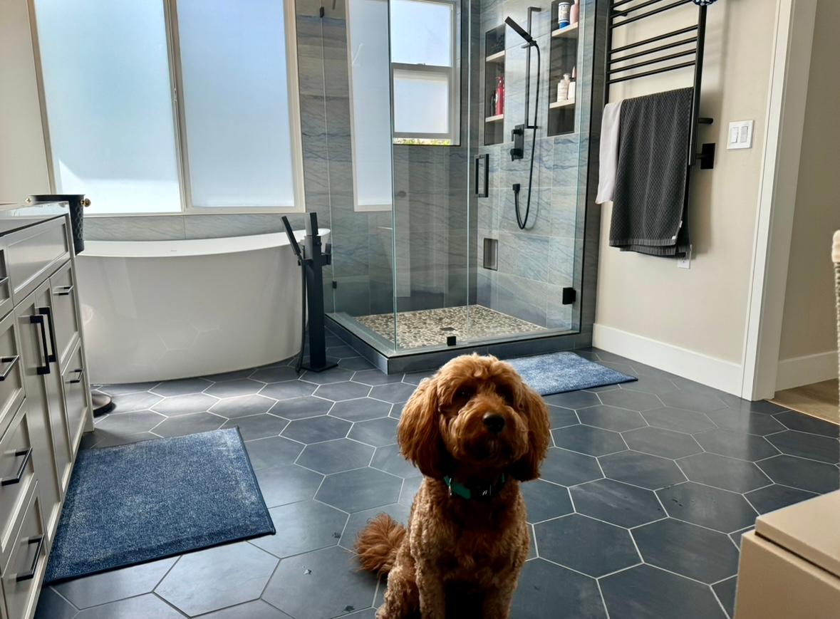 A dog is sitting in a bathroom next to a bathtub and shower.