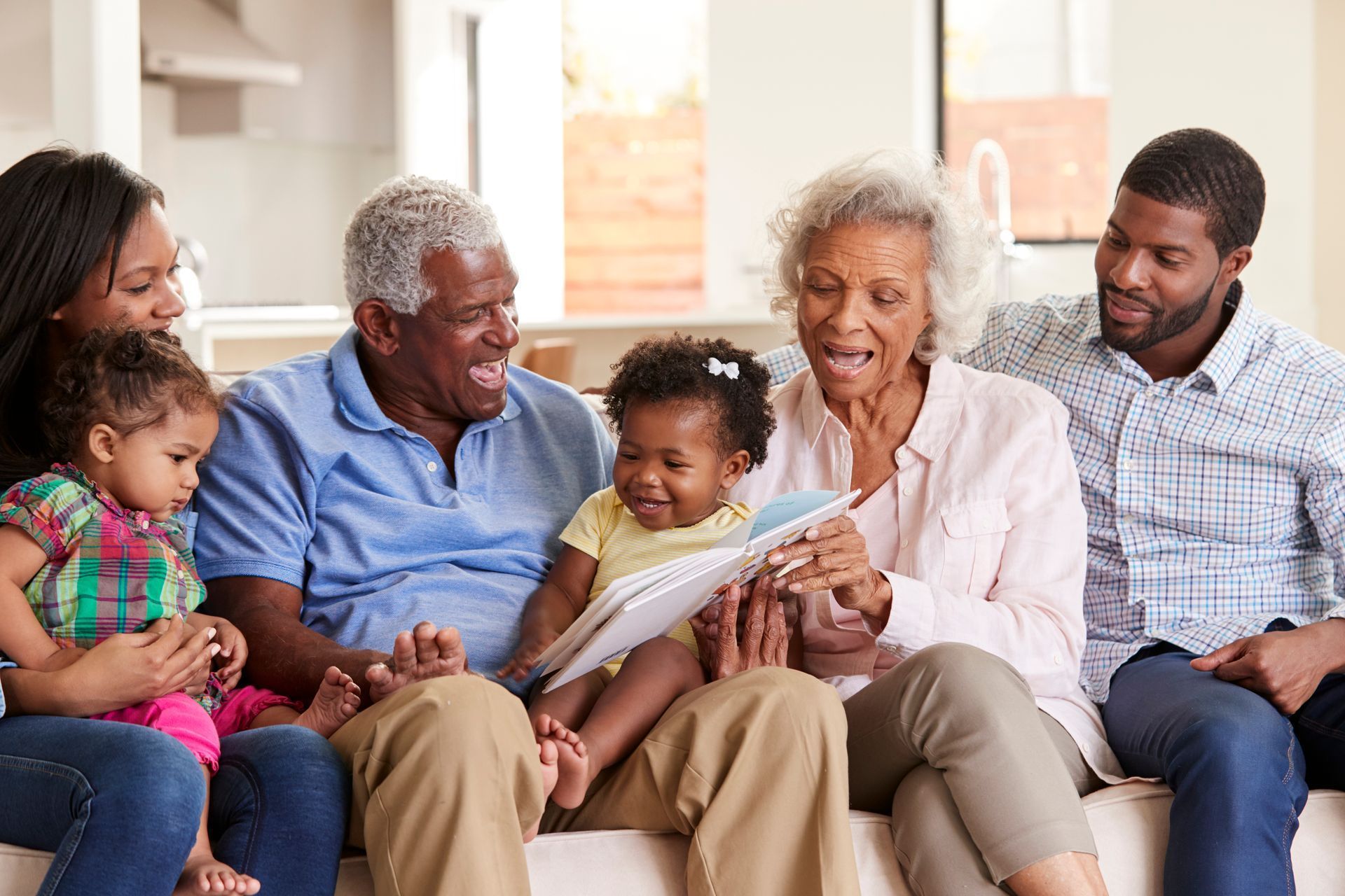 Multi-generational family smiling while reading a book together on a sofa in a brightly lit living room