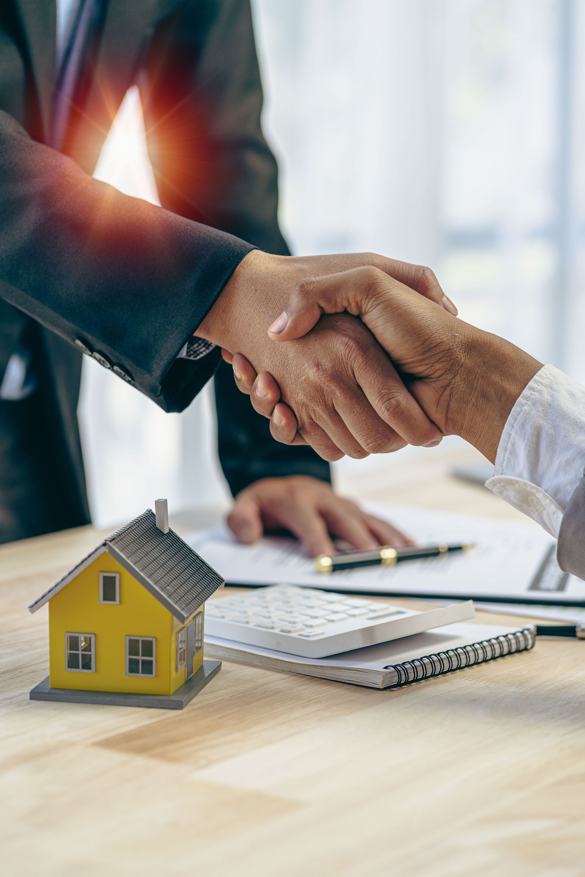 Two people shaking hands over a table with a model house, papers, and a calculator.