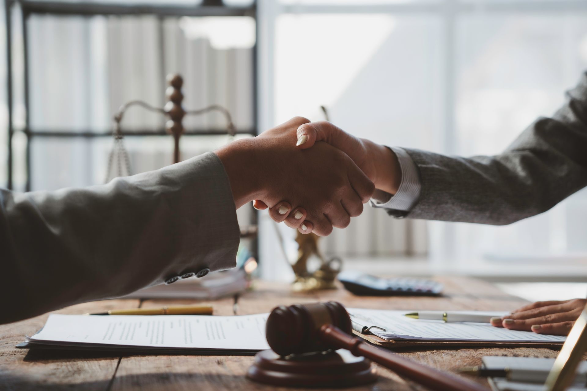 Two people in suits shaking hands over a wooden table with a gavel, scales of justice, and documents.