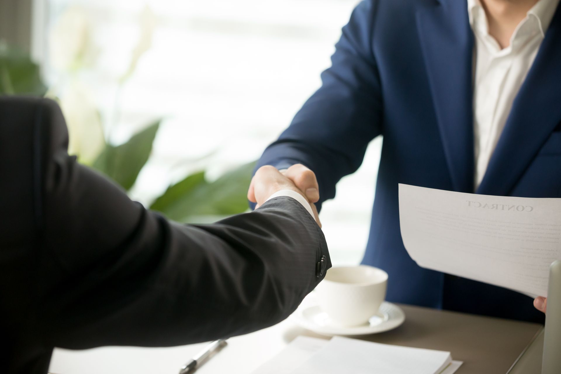 Two men in suits shaking hands over a table with a coffee cup and papers, indoors.