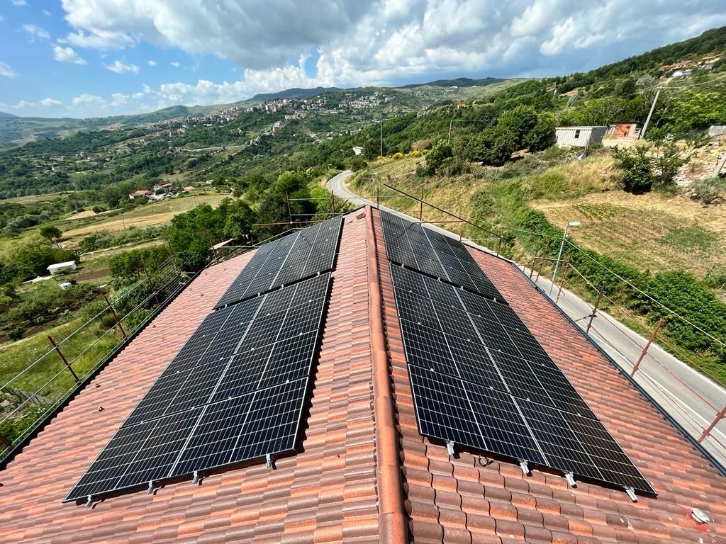 Pannelli solari su un tetto di tegole rosse che si affacciano su una verde collina sotto un cielo nuvoloso.