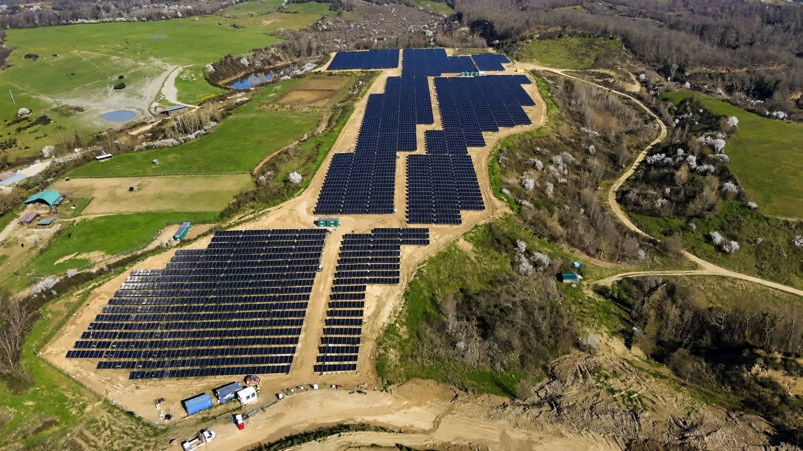 Veduta aerea di un grande impianto solare situato su una collina, tra campi verdi e terreno roccioso.