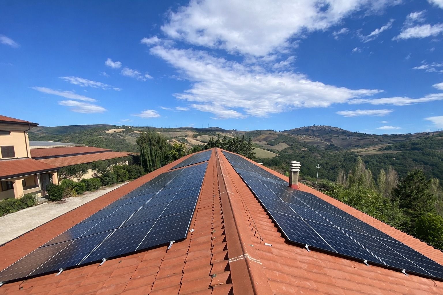 Pannelli solari installati lungo il colmo di un tetto in terracotta con vista sulle colline e sul cielo azzurro.