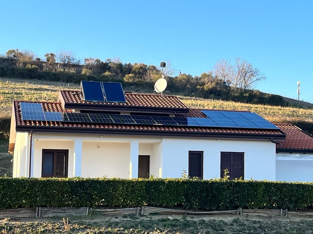 Casa bianca con pannelli solari su una collina soleggiata, con una siepe e il cielo azzurro sullo sfondo.