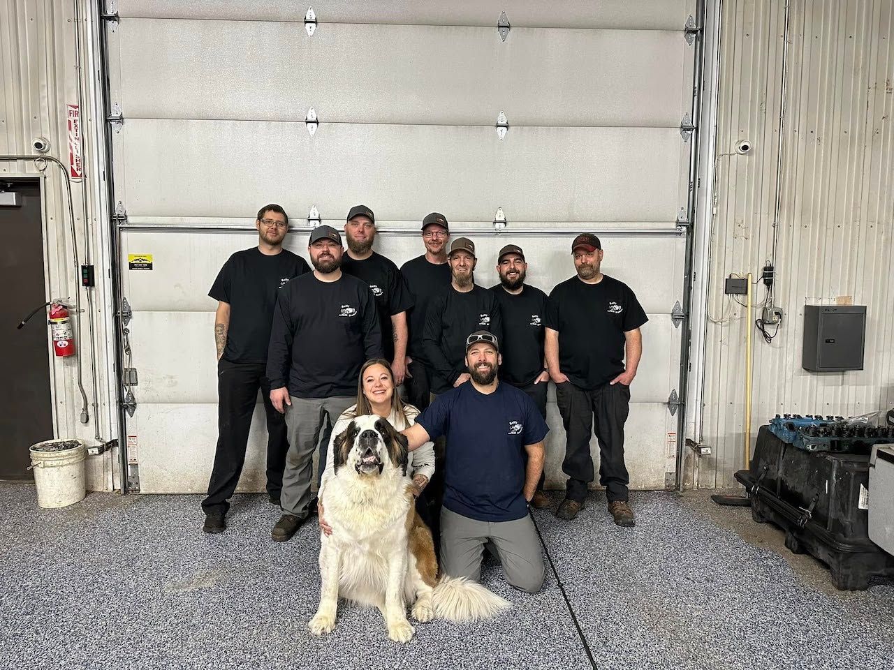 Six people and a dog posing in a warehouse with a large white garage door. | Belly Brothers Auto Tech