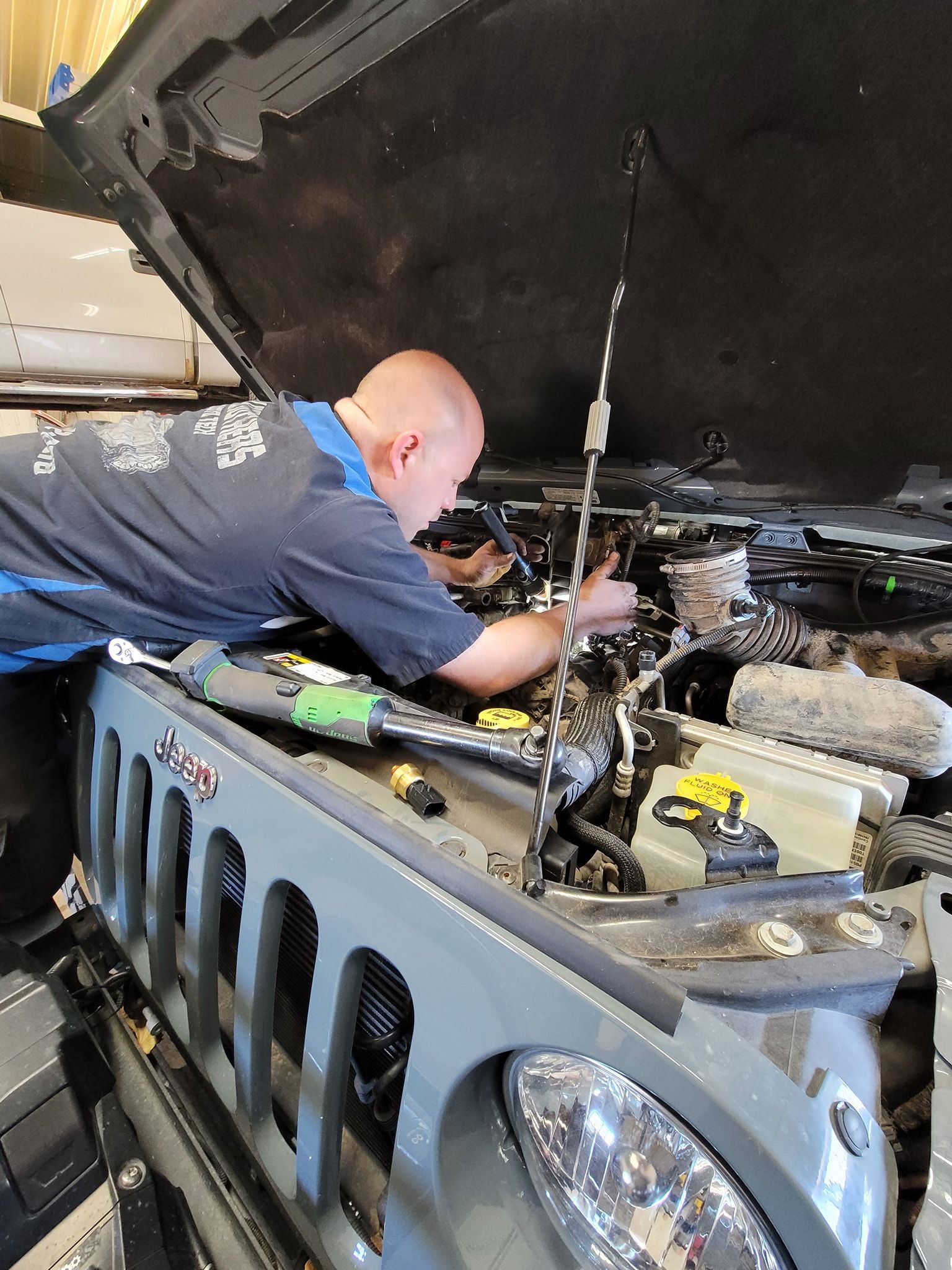Mechanic working under the open hood of a gray Jeep in a garage 