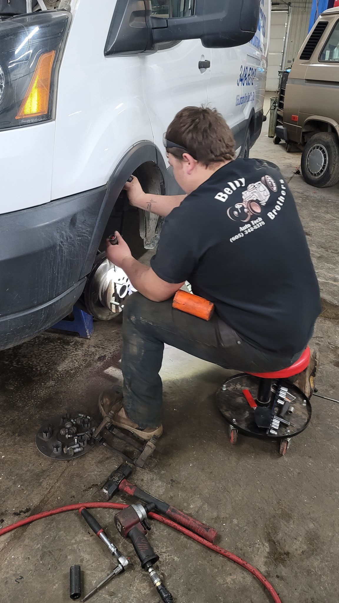 Mechanic crouches beside a white van, working on the front wheel in a garage. | Belly Brothers Auto Tech