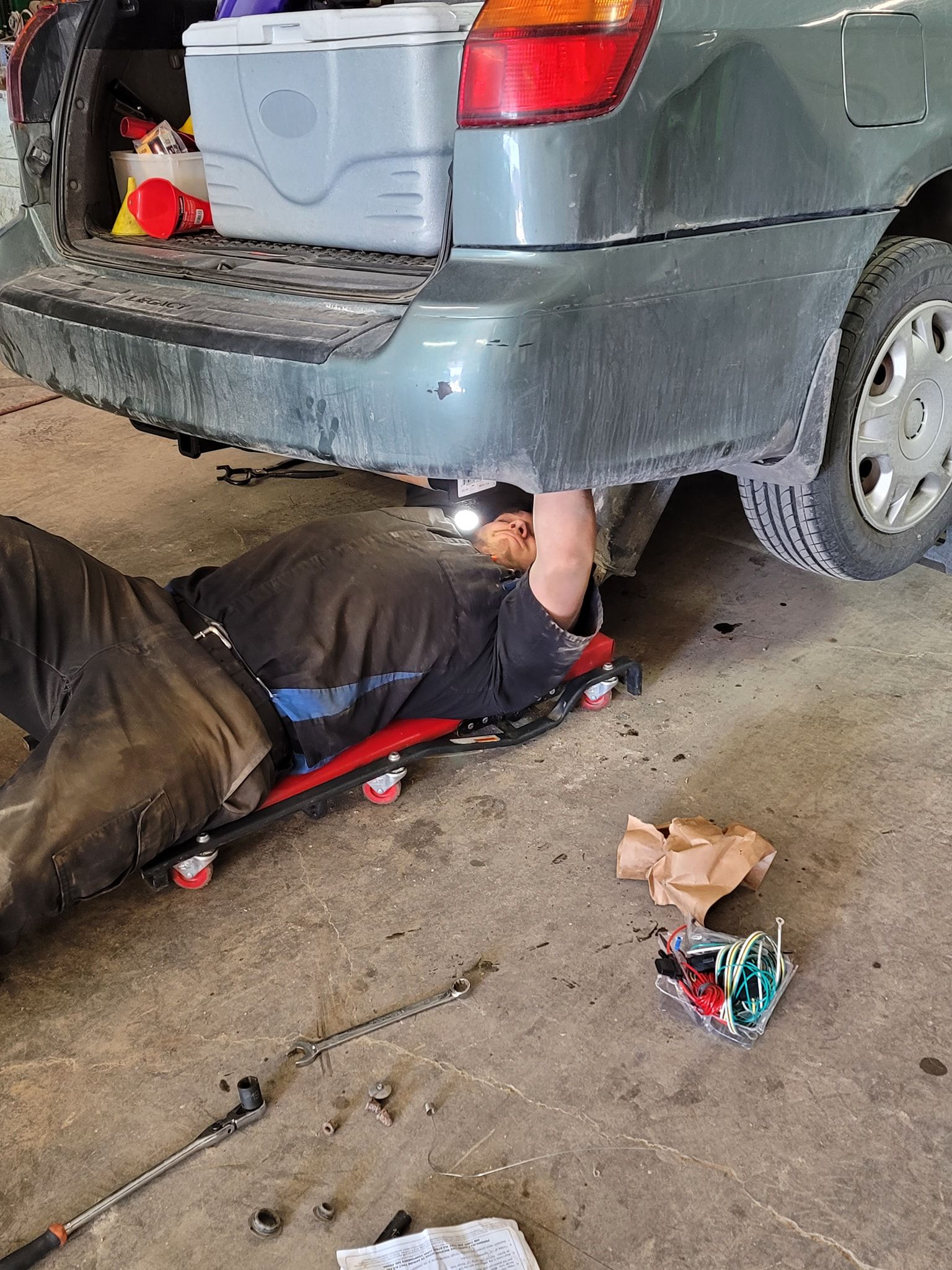 Mechanic lying under a gray SUV, working on the rear underside in a garage. | Belly Brothers Auto Tech