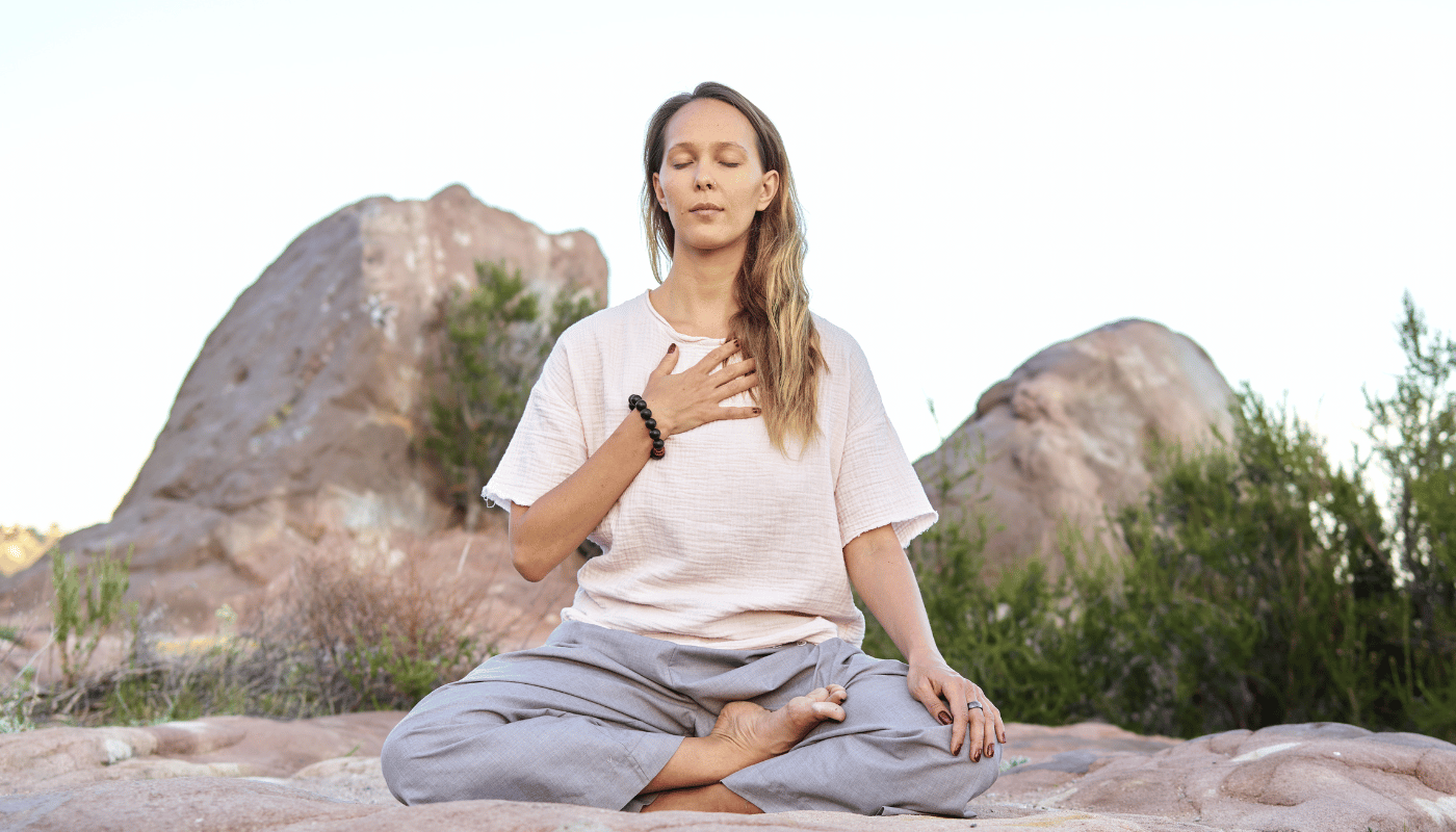 A woman is sitting in a lotus position on a rock with her eyes closed.